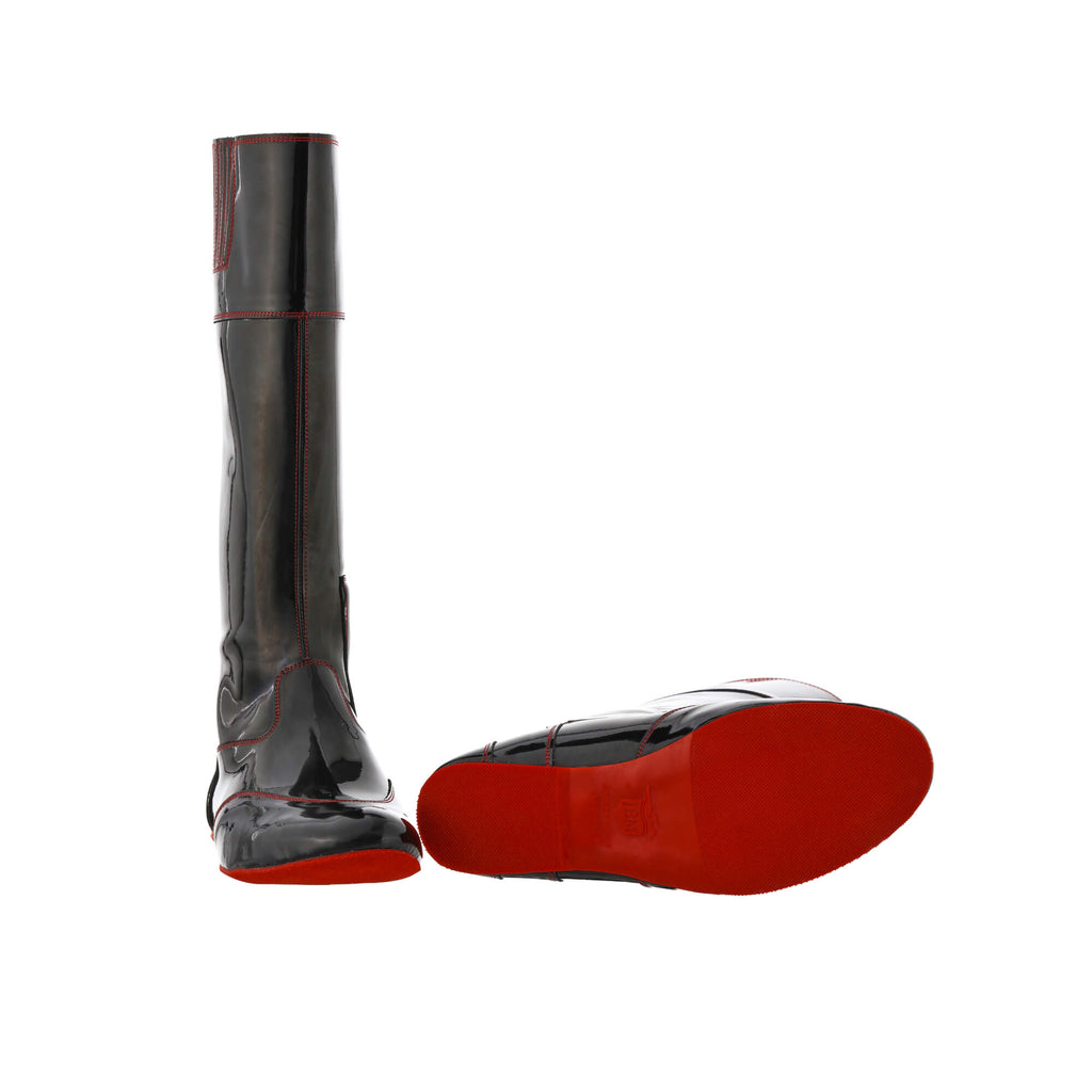 A pair of tall racing boots displayed against a white background. The boots are made of black material with red exterior stitching and red soles. They feature a back zip and a 1.5-2cm stretch feature around the calf. One boot is positioned sole down with the zipper done, while the other boot is laid down to showcase the sole.