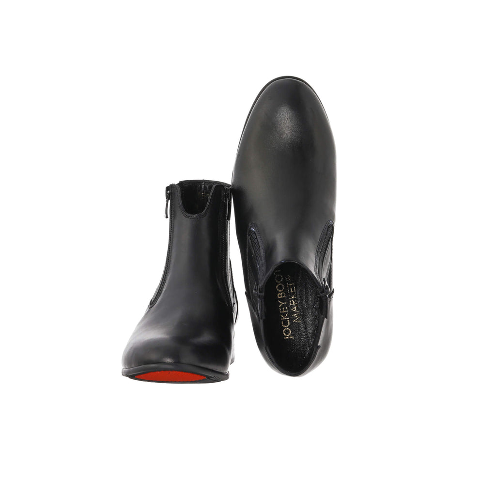 A pair of classic black short boots featuring double front zip and a gap on the top front to provide extra comfort when horse riding. Presented on a white background. The first shoe's sole is placed on the floor, and the zips are done. The second shoe is upside down with the sole facing behind, showcasing the inside of the shoe.