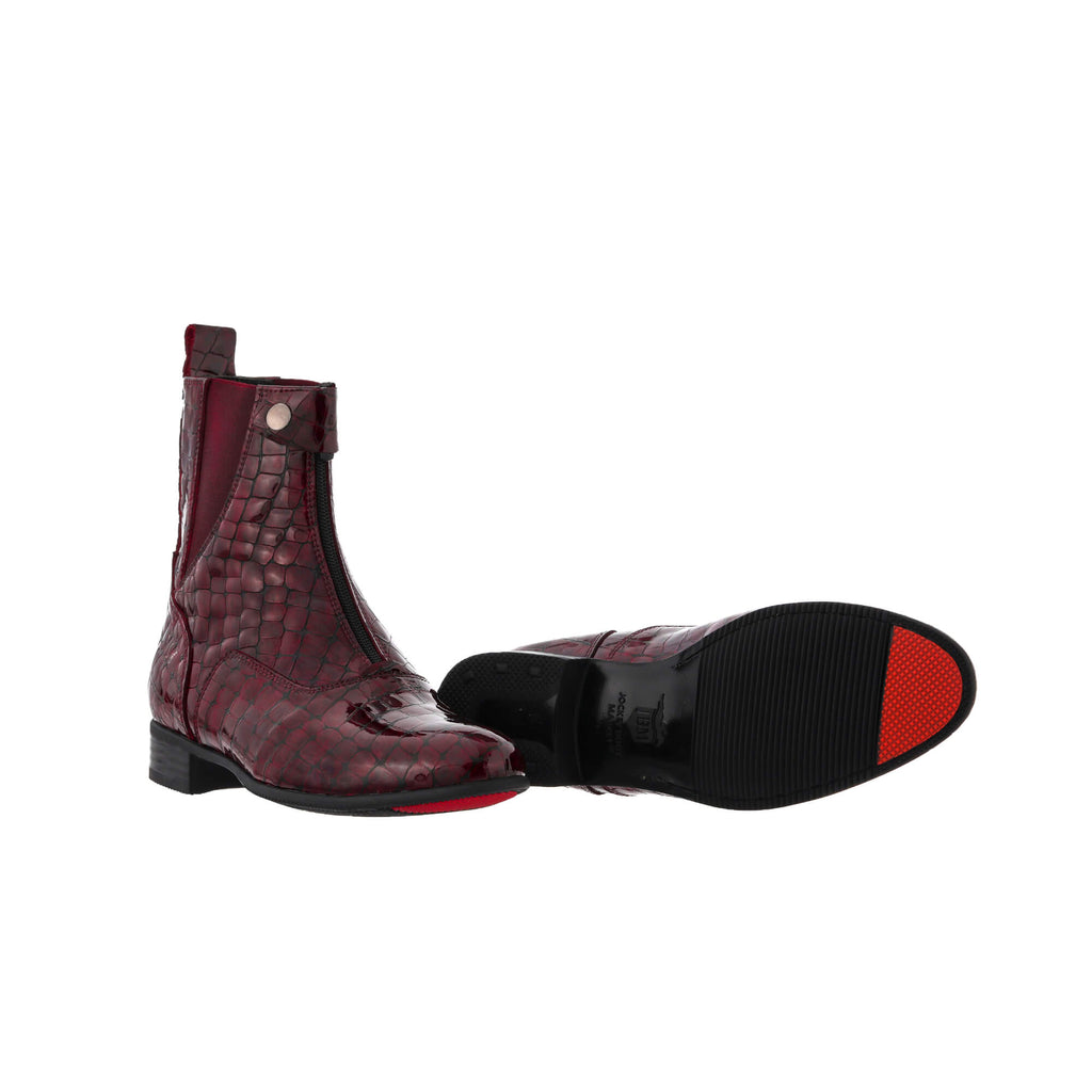 A pair of burgundy crocodile leather boots displayed against a white background. One boot is positioned sole-down with the buckle and front zip closure completed. The other boot is laid flat to showcase its sole, featuring a red-tipped signature design.